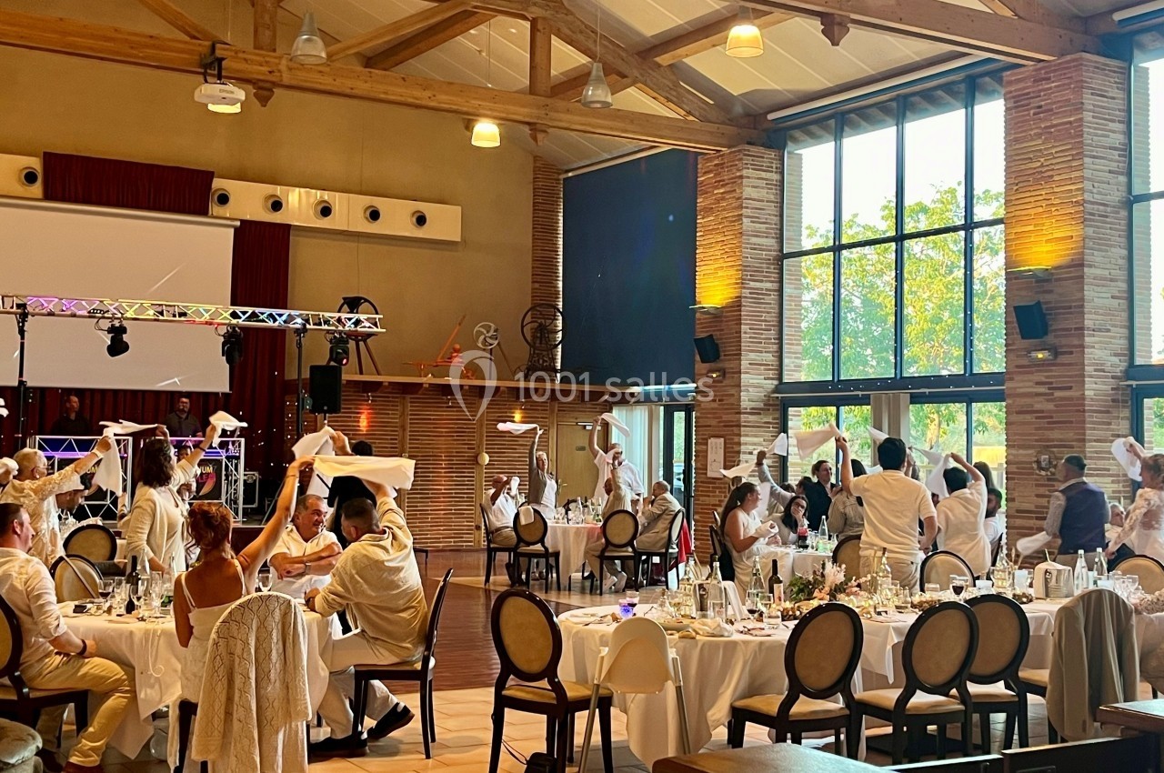 Salle de réception lumineuse avec des invités en tenue blanche célébrant autour de tables décorées.