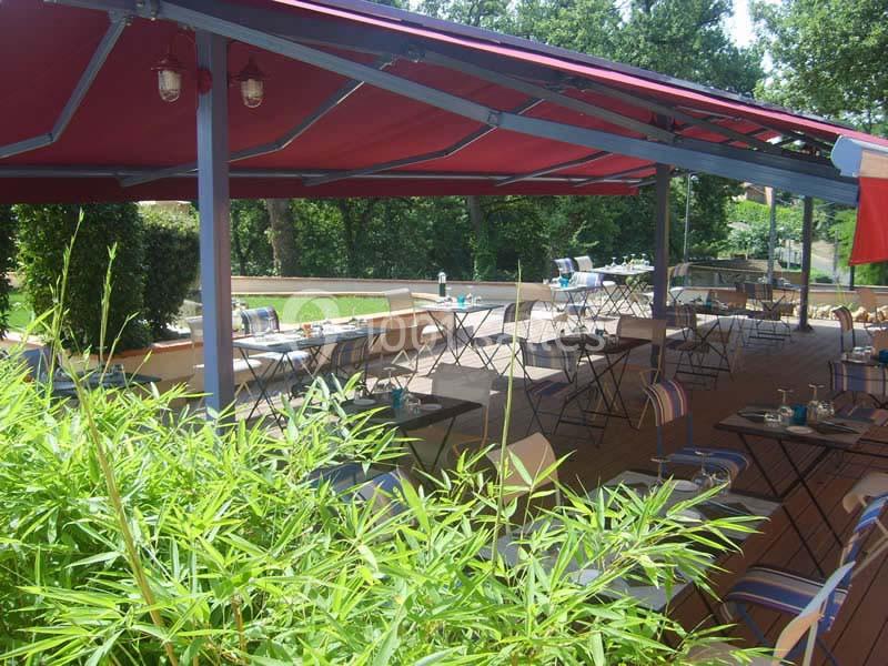 Terrasse extérieure ombragée avec tables et chaises, entourée de verdure et protégée par une pergola rouge.