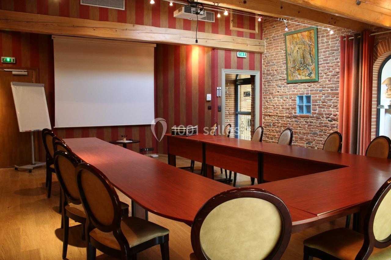 Salle de réunion avec tables en bois disposées en U, chaises rembourrées, écran de projection et mur en briques apparentes.