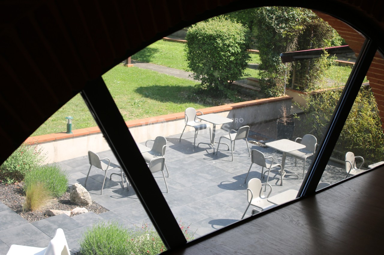Vue d'une terrasse extérieure avec des tables et des chaises, entourée de verdure, prise depuis une fenêtre en arc.