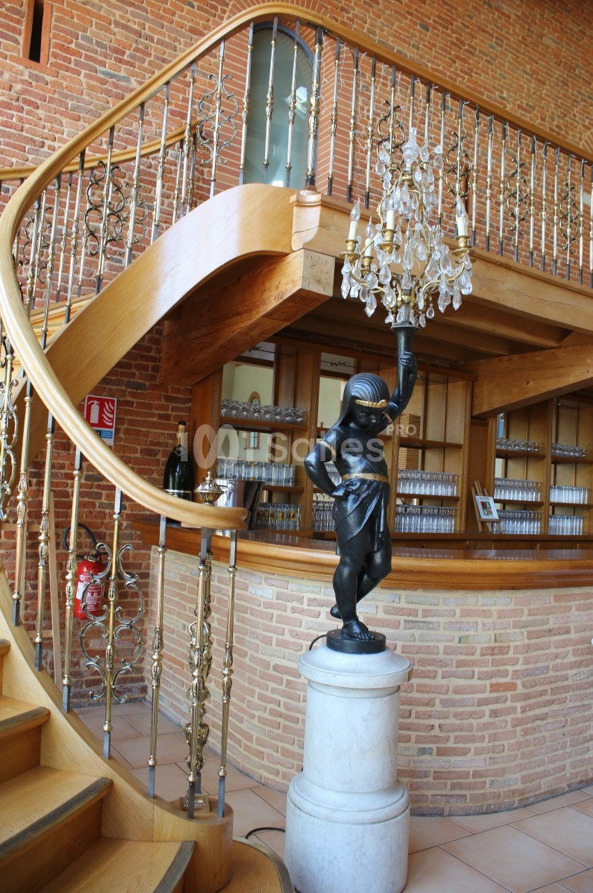 Escalier en bois courbé avec balustrade dorée, statue décorative sur socle et lustre suspendu devant un mur en briques.