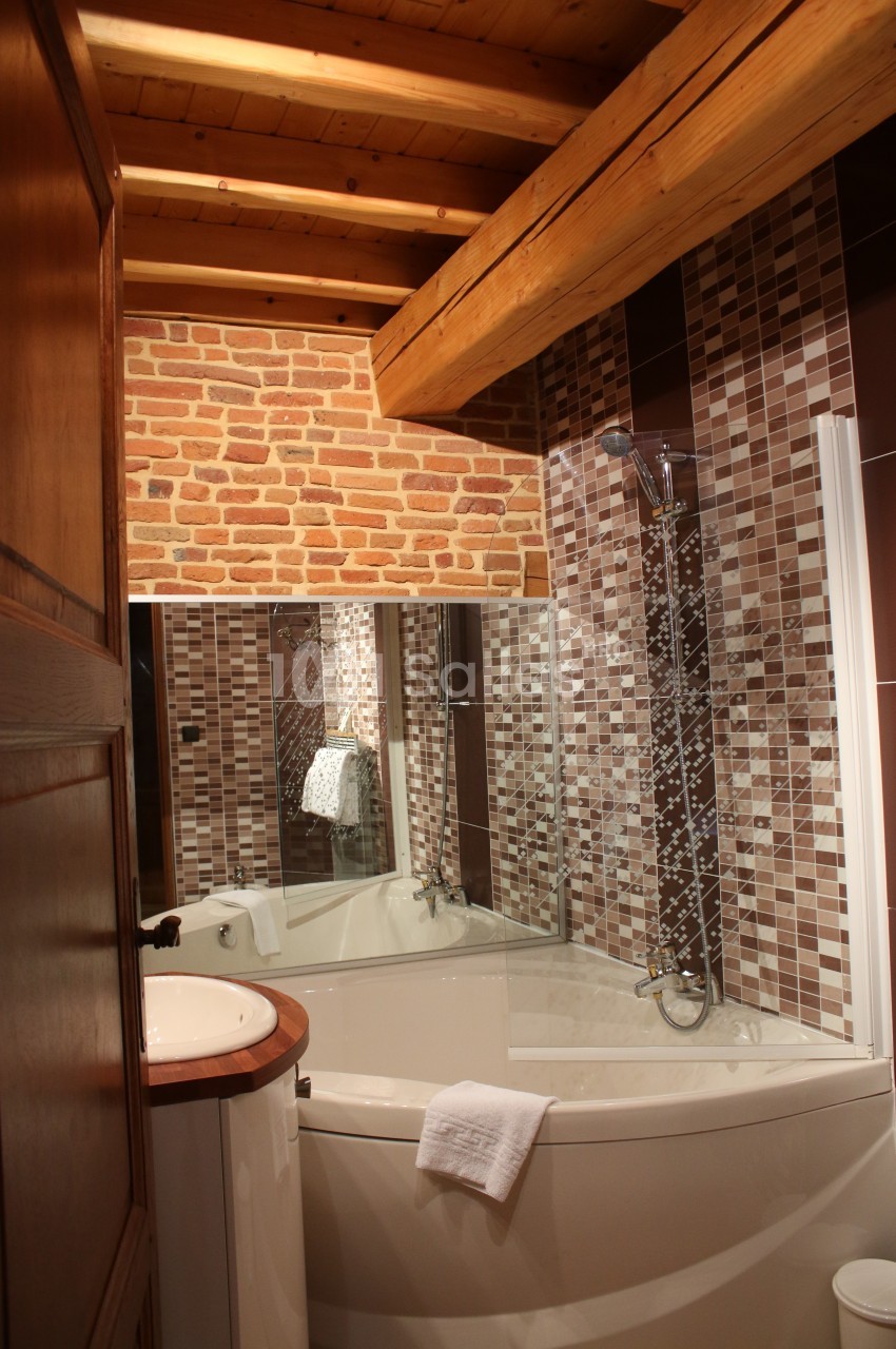 Salle de bain avec baignoire d'angle, mur en briques et carrelage mosaïque, sous un plafond en bois.