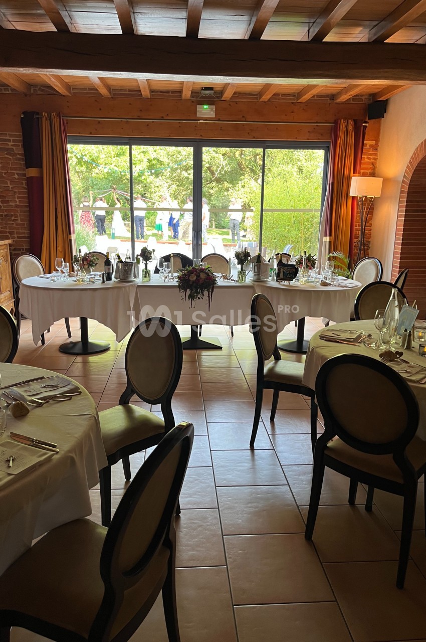 Salle de réception lumineuse avec tables rondes dressées, vue sur un jardin à travers de grandes baies vitrées.