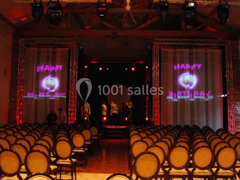 Salle de réception avec des rangées de chaises vides, scène éclairée et écrans affichant ’Happy Birthday’.