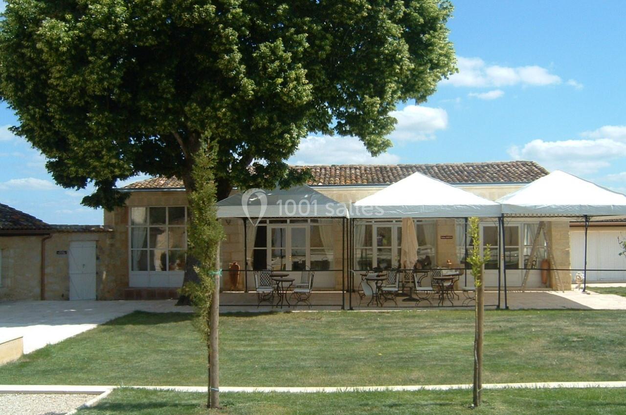 Maison en pierre avec terrasse ombragée par des tonnelles blanches, entourée d'un jardin et d'un grand arbre.