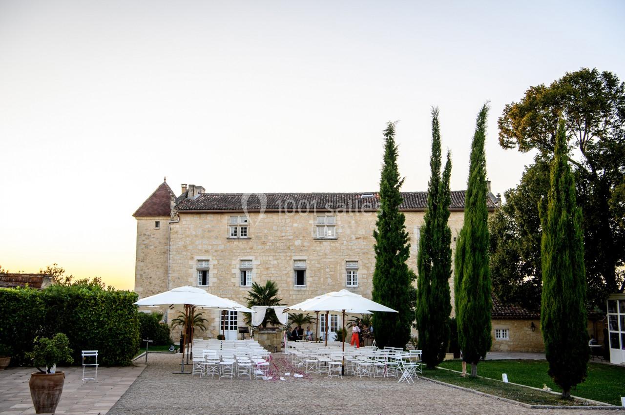 Cour d'un château en pierre avec des tables et chaises blanches disposées sous des parasols, entourée de cyprès.