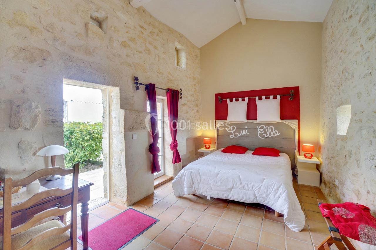 Chambre lumineuse avec murs en pierre, lit double, rideaux rouges et accès à une terrasse extérieure.