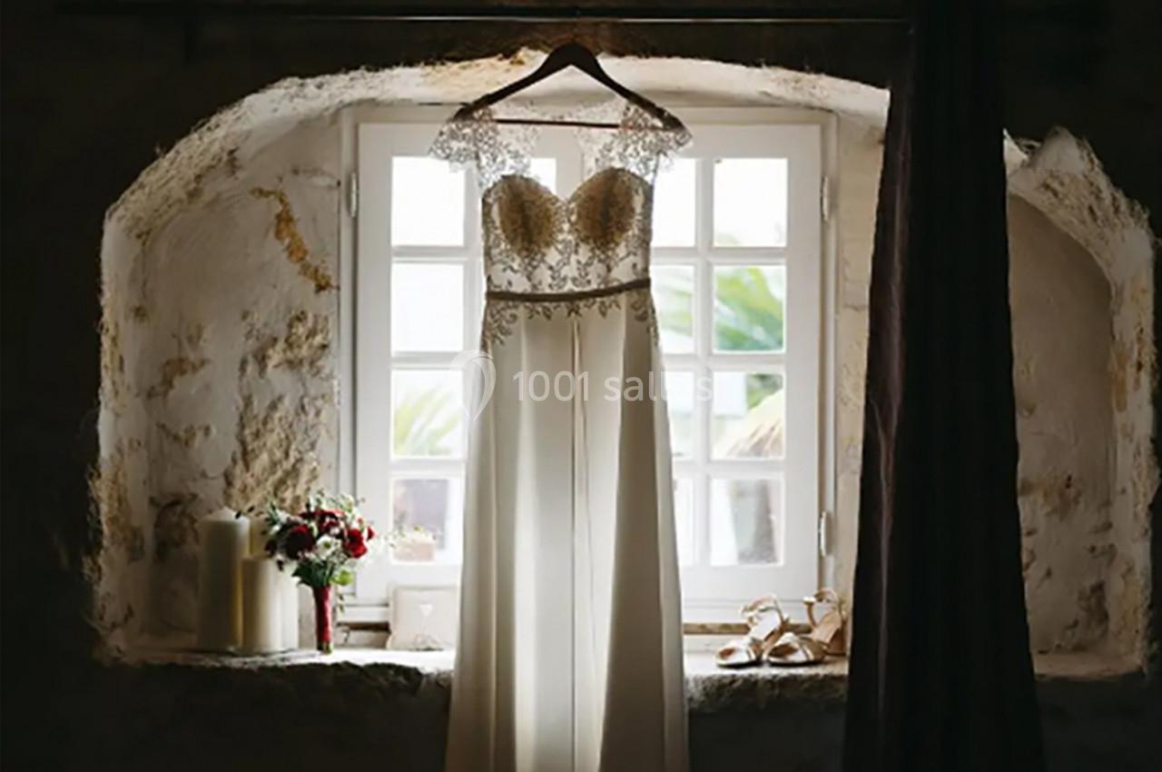 Robe de mariée suspendue devant une fenêtre lumineuse, avec des bougies, un bouquet et des chaussures posés en dessous.