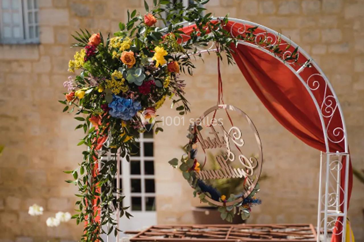 Arche décorative en métal blanc ornée de fleurs colorées et de rubans rouges, devant un mur en pierre beige.