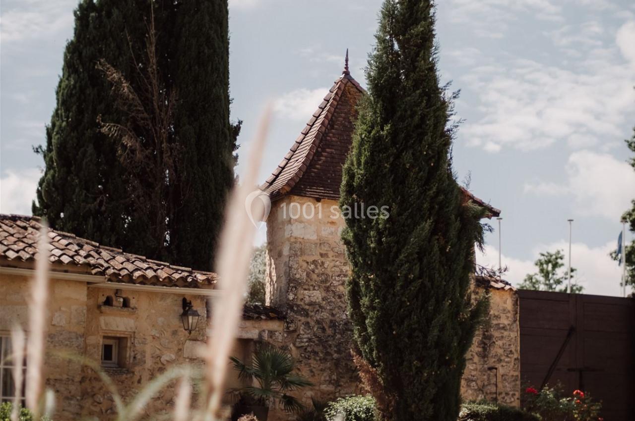 Vue d'un jardin avec des cyprès, un bâtiment en pierre aux tuiles rouges et un ciel partiellement nuageux.