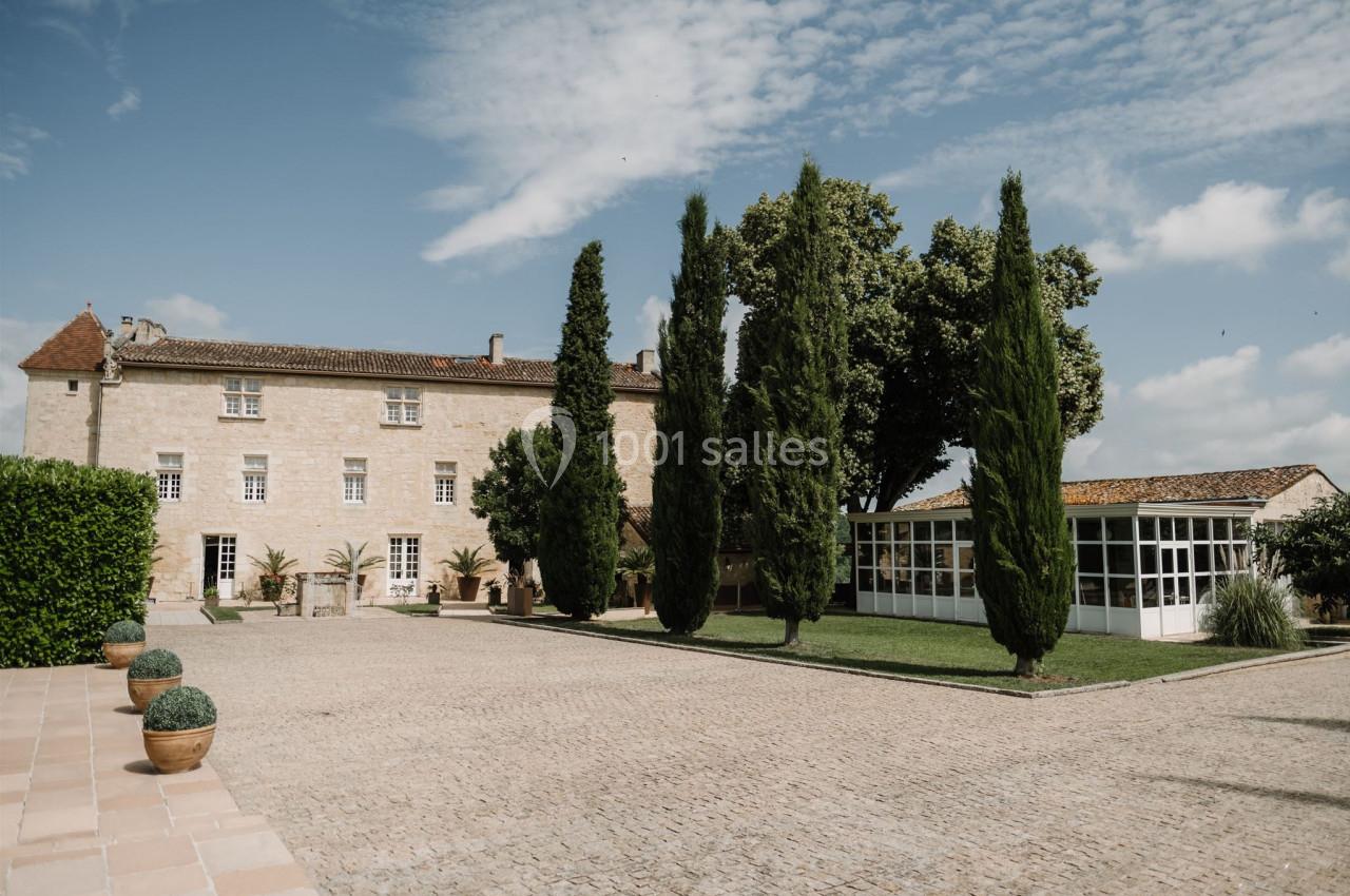 Cour pavée avec un bâtiment en pierre, des cyprès et une véranda entourés de verdure sous un ciel partiellement nuageux.