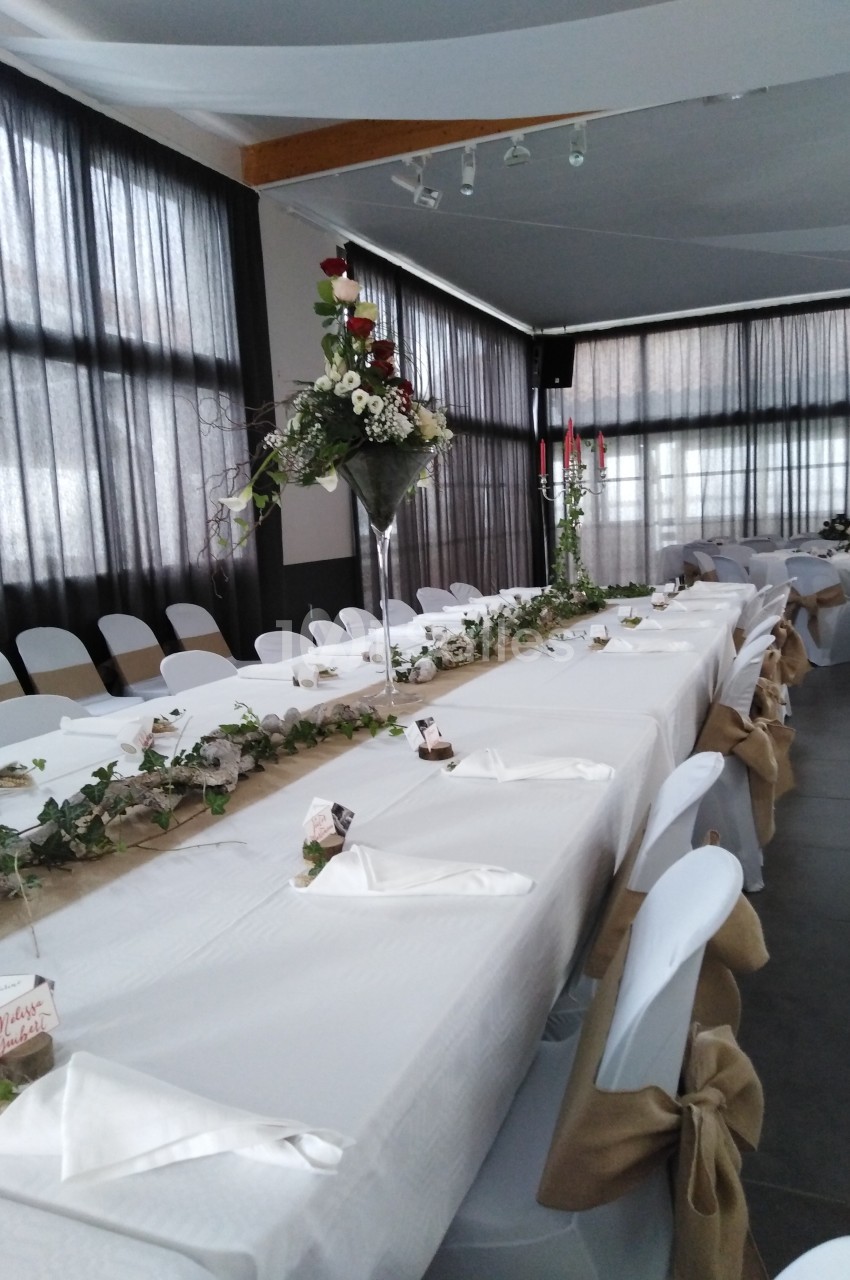 Salle décorée pour un événement avec une longue table dressée, nappes blanches, chaises ornées et centres de table fleuris.