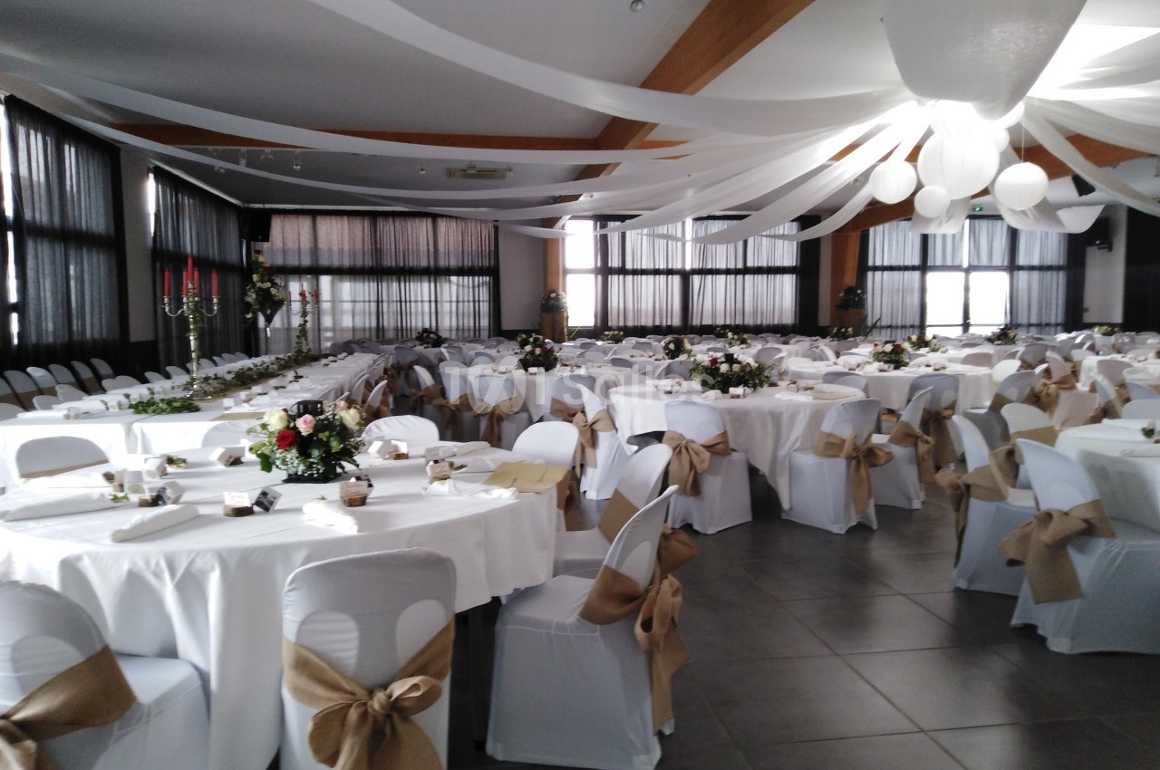 Salle de réception décorée avec des tables rondes, nappes blanches, chaises ornées de nœuds en toile de jute et guirlandes…