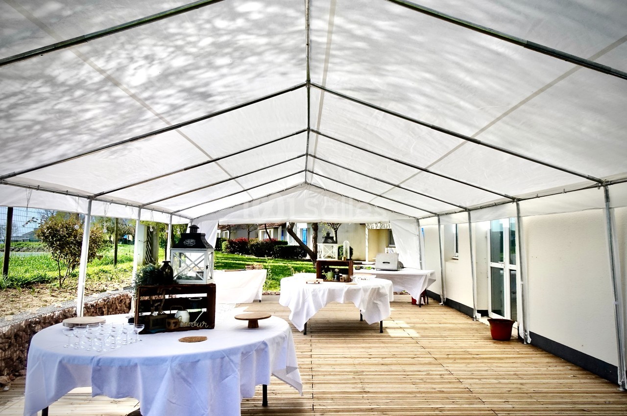 Tente blanche installée sur une terrasse en bois, avec des tables rondes et rectangulaires dressées pour un événement.
