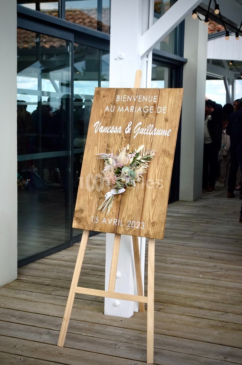 Panneau en bois sur un chevalet annonçant un mariage, décoré de fleurs séchées, placé sur une terrasse en bois.