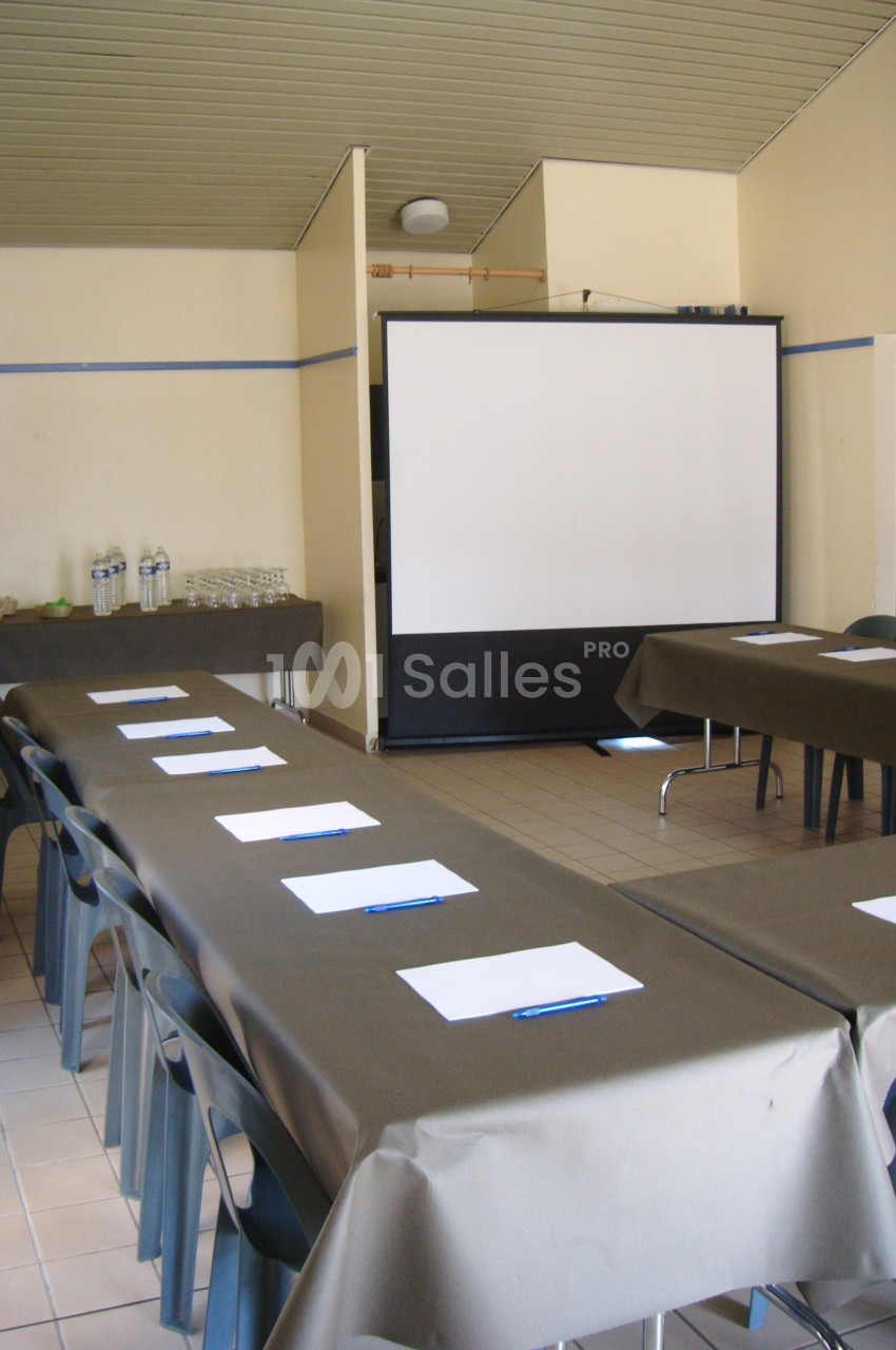 Salle de réunion avec tables disposées en U, nappes brunes, chaises, écran de projection et fournitures de bureau.