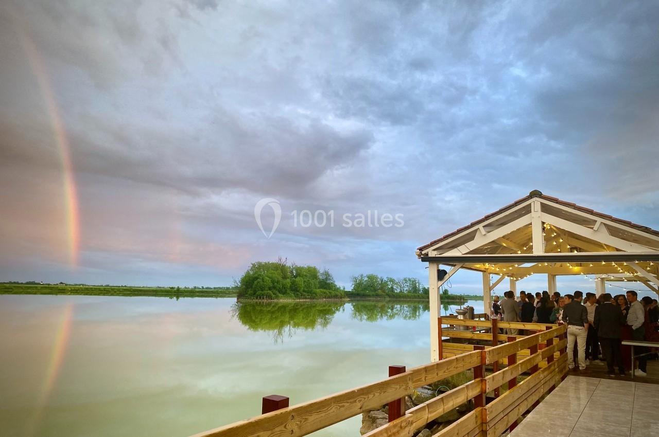 Un groupe de personnes rassemblé sous une terrasse éclairée, au bord d'un lac avec un arc-en-ciel visible dans le ciel.
