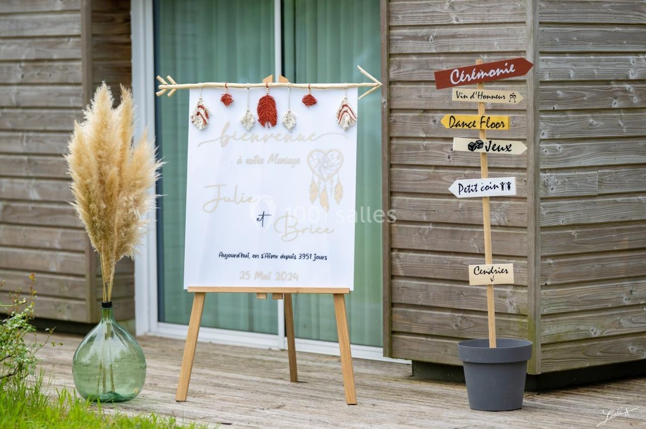 BIENVENUE AUX INVITES Panneau de bienvenue pour un mariage devant un bâtiment en bois, avec décorations et pancartes directionnelles.