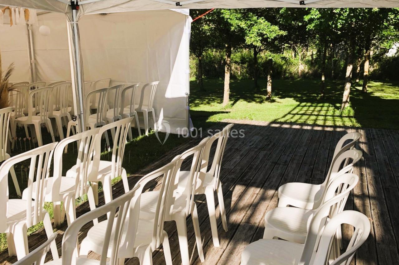CEREMONIE LAIQUE Chaises blanches en plastique alignées sous une tente blanche sur une terrasse en bois, avec un jardin en arrière-plan.