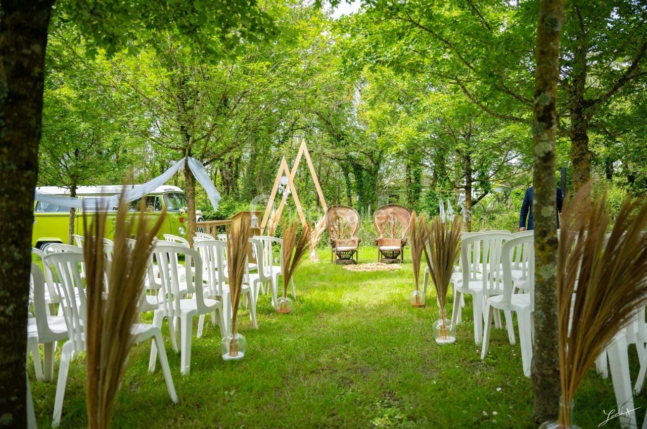 CEREMONIE LAIQUE Allée de chaises blanches disposées en extérieur, menant à une arche en bois et des fauteuils sous des arbres.