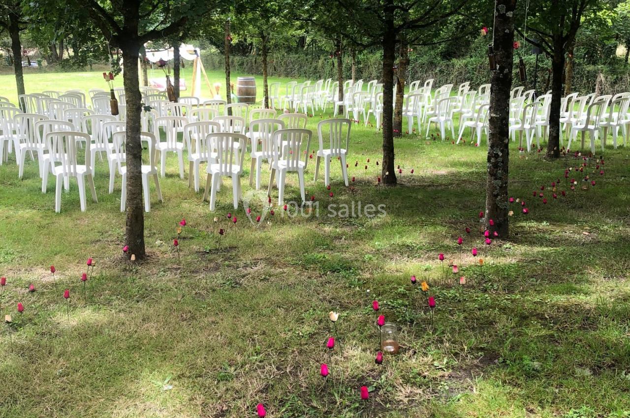 CEREMONIE LAIQUE Chaises blanches disposées en rangées sous des arbres, avec des fleurs rouges plantées dans l'herbe.