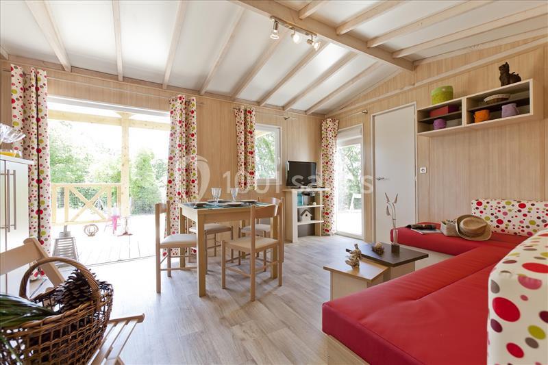 CHALET 2 CHAMBRES Salon lumineux avec canapé rouge, table à manger en bois, étagères colorées et vue sur une terrasse extérieure verdoyante.