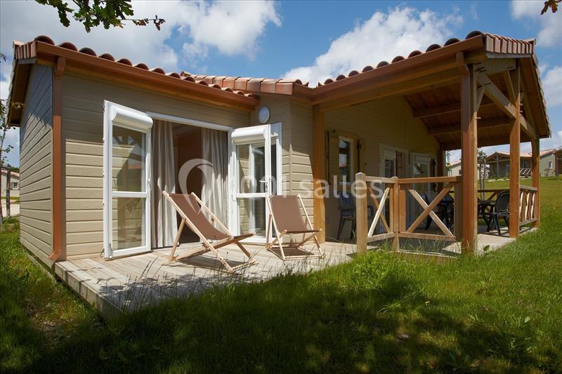 CHALETS 3 CHAMBRES Chalet en bois avec terrasse, deux chaises longues et mobilier de jardin, entouré de pelouse sous un ciel partiellement…