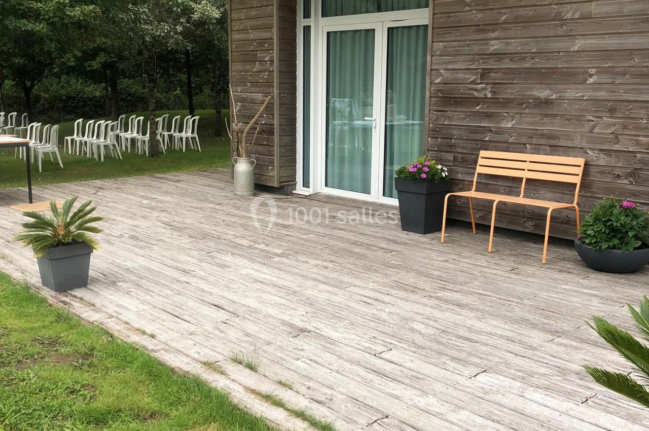 L'ESPACE DU LAC Terrasse en bois avec banc, pots de fleurs et pergola, donnant sur une pelouse et des chaises blanches en arrière-plan.