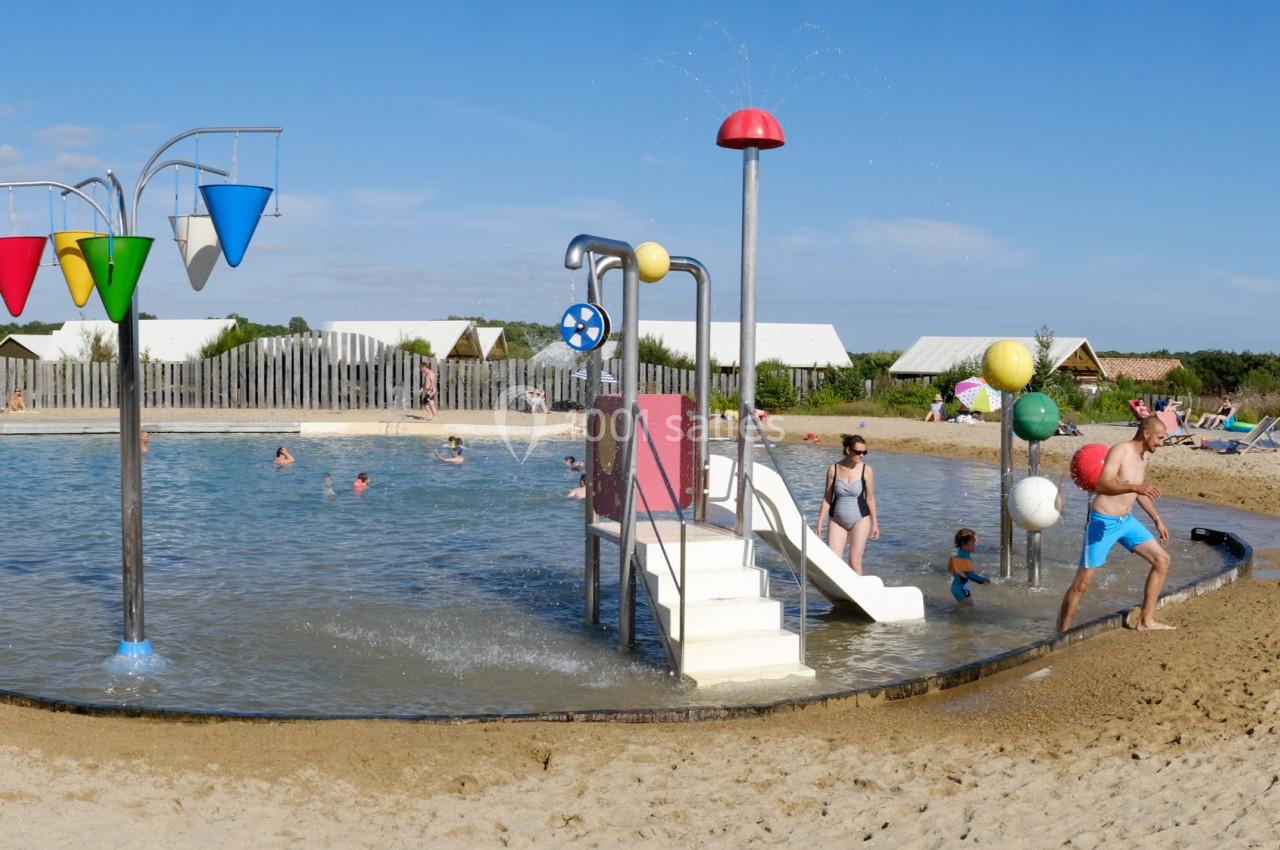 Lac de baignade Bassin peu profond avec jeux d'eau colorés, toboggan et plage de sable, entouré de végétation et d'une clôture en bois.