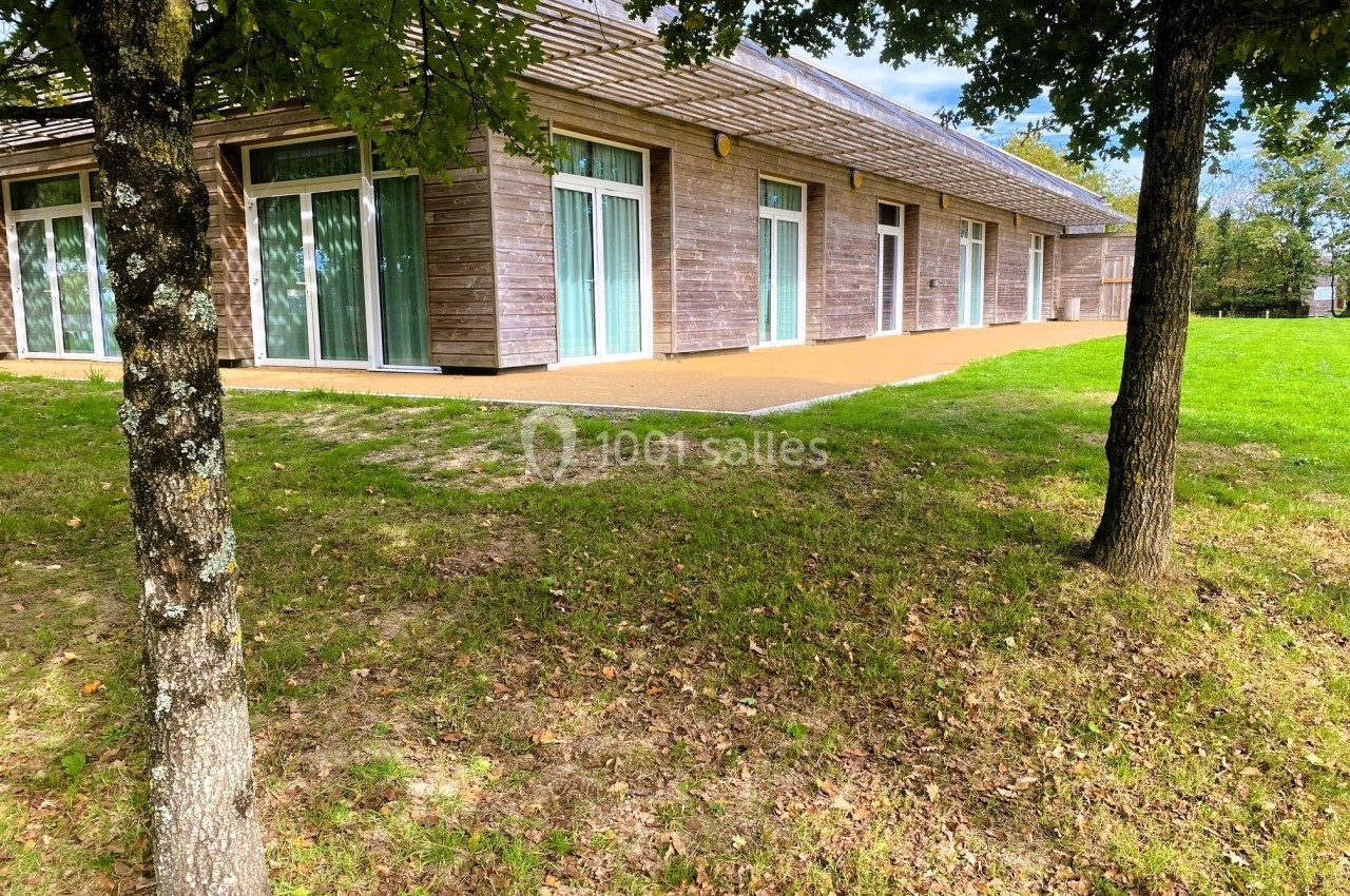 NOUVELLE TERRASSE Bâtiment en bois avec de grandes baies vitrées, entouré d'herbe et d'arbres sous un ciel partiellement dégagé.