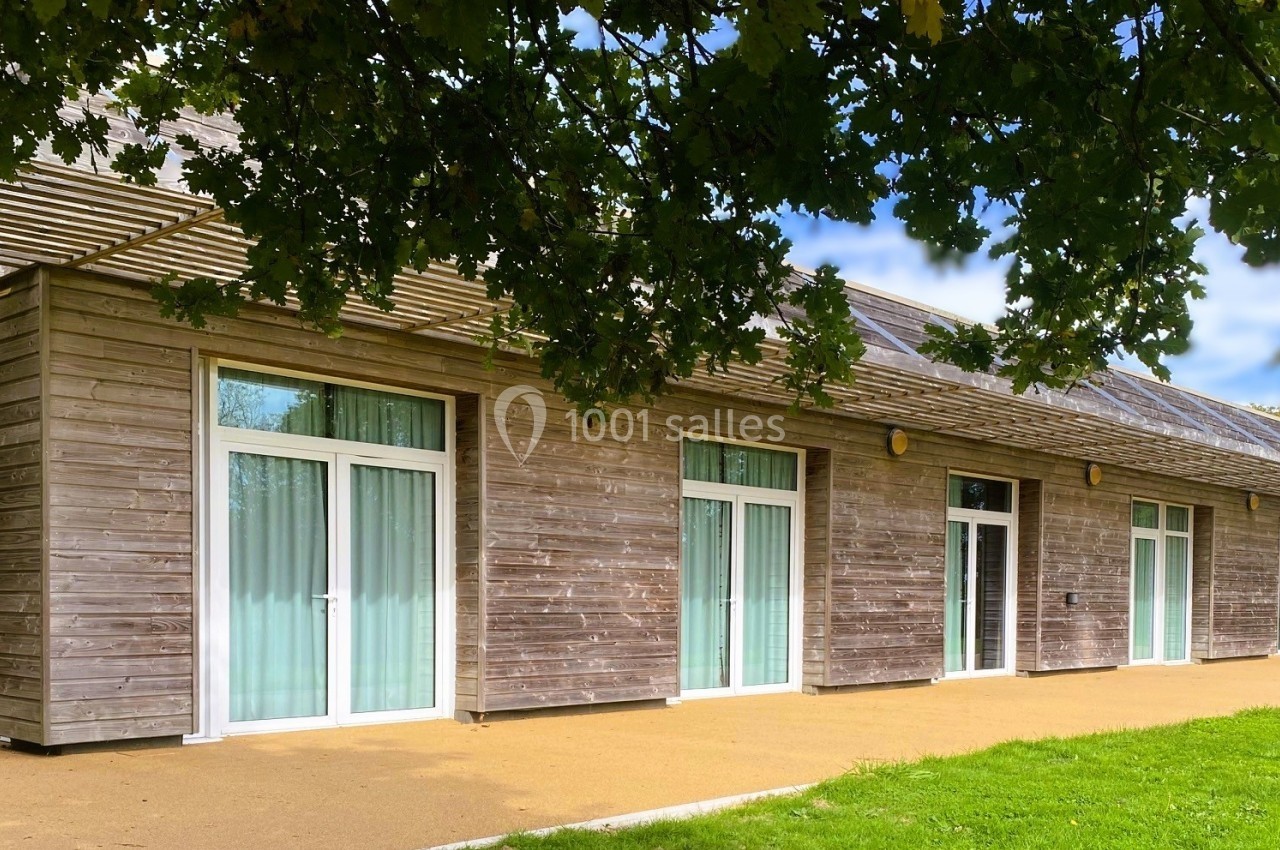 NOUVELLE TERRASSE Bâtiment en bois avec de grandes baies vitrées, entouré de verdure et partiellement ombragé par des arbres.
