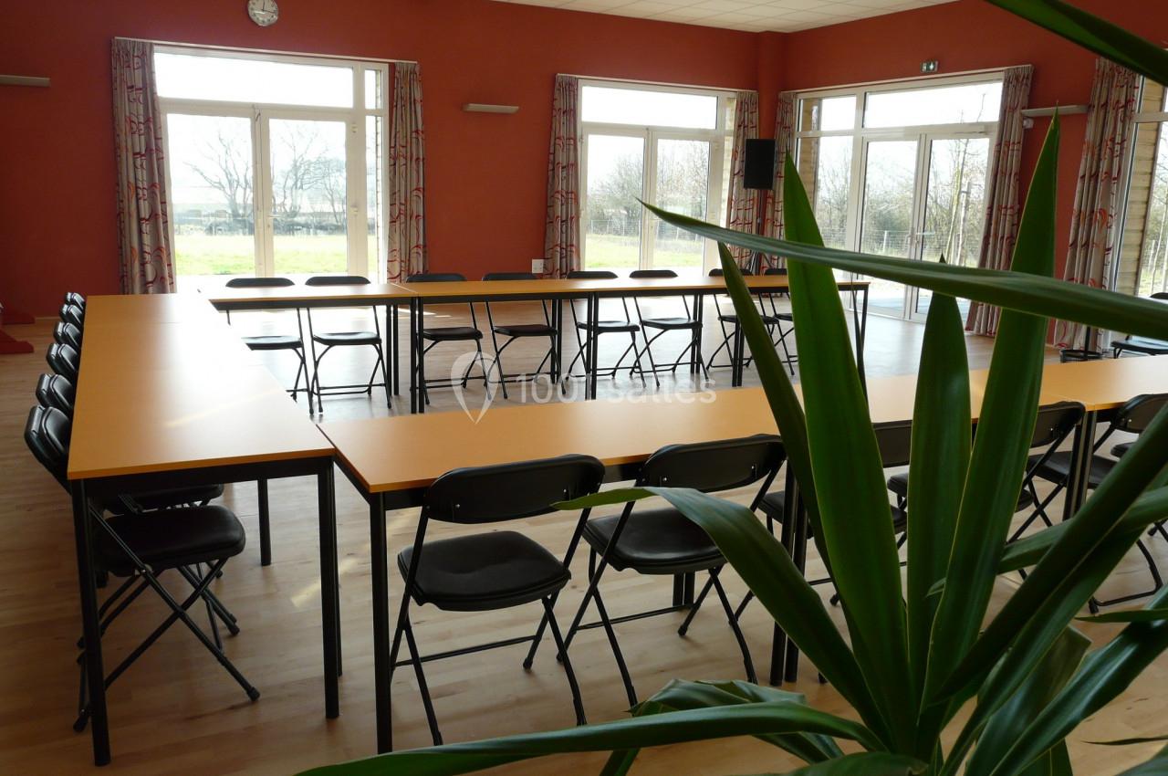 SALLE BELLEVUE SEMINAIRE Salle lumineuse avec des tables disposées en U, des chaises pliantes noires et une plante au premier plan.
