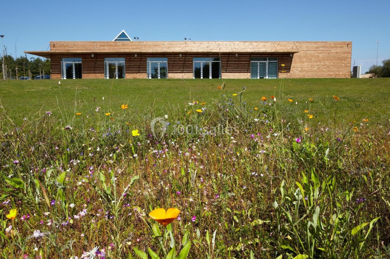 SALLE DE RECEPTION Bâtiment en bois avec de grandes baies vitrées, entouré d'une prairie fleurie sous un ciel bleu.