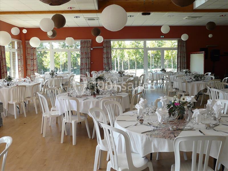 SALLE LE JAUNAY Salle décorée pour un événement, avec des tables rondes dressées, des chaises blanches et des lanternes suspendues.