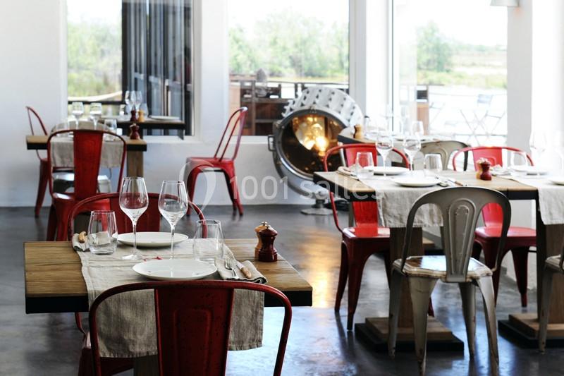 Salle de restaurant lumineuse avec des tables en bois dressées, chaises rouges et blanches, et grandes fenêtres ouvertes.