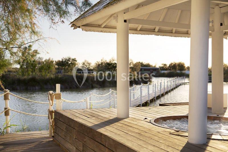 Terrasse en bois avec un jacuzzi sous un kiosque, vue sur un ponton bordant un plan d'eau entouré de végétation.