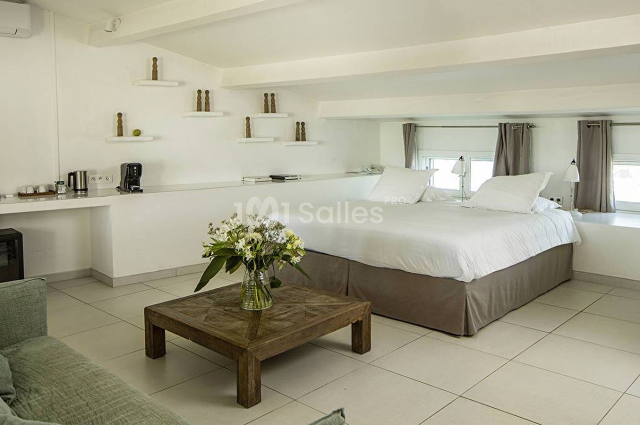 Chambre lumineuse avec lit double, table basse en bois, canapé vert et étagères décoratives sur un mur blanc.