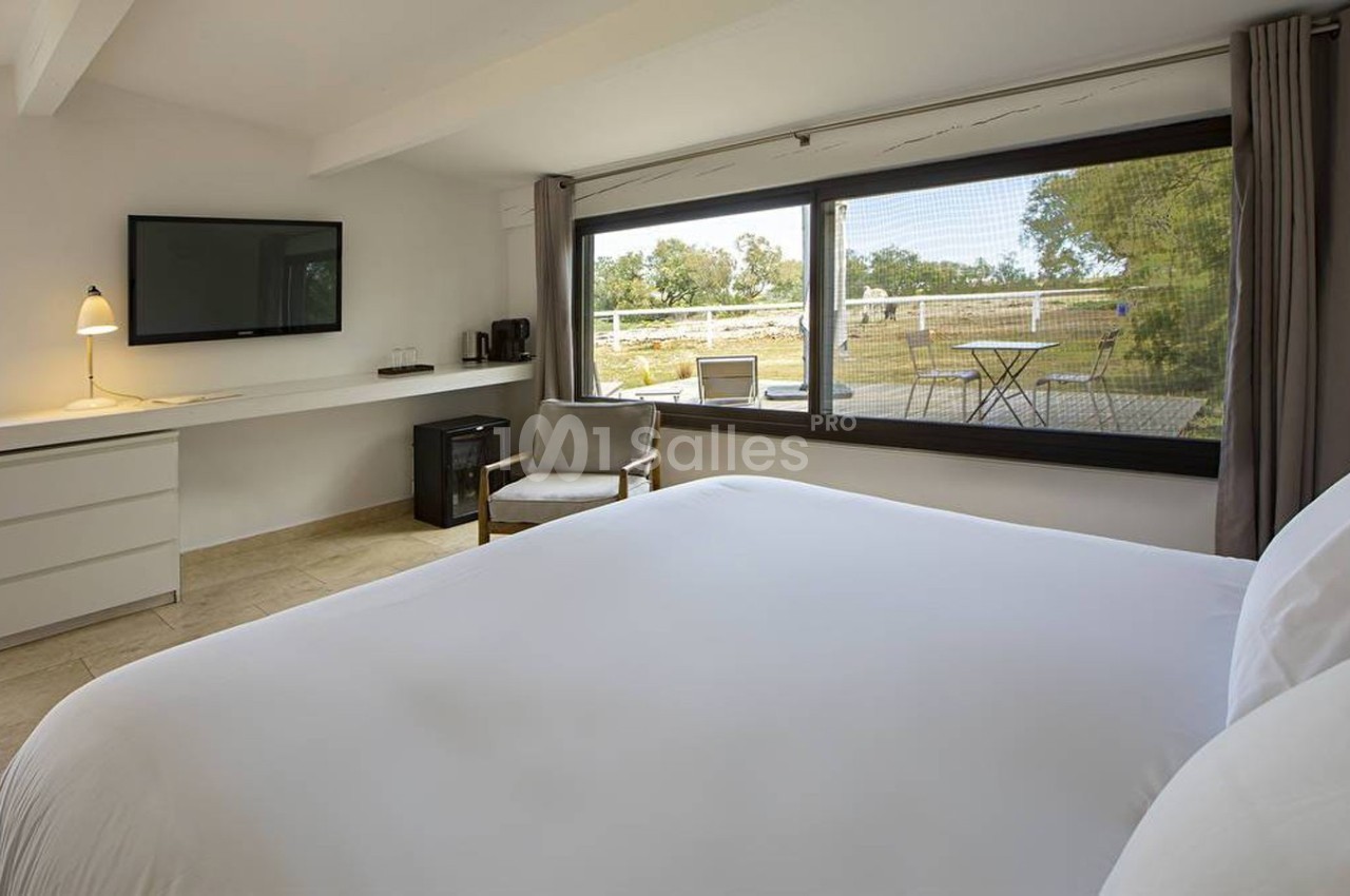Chambre lumineuse avec lit double, bureau, télévision, fauteuil et grande baie vitrée donnant sur un jardin.