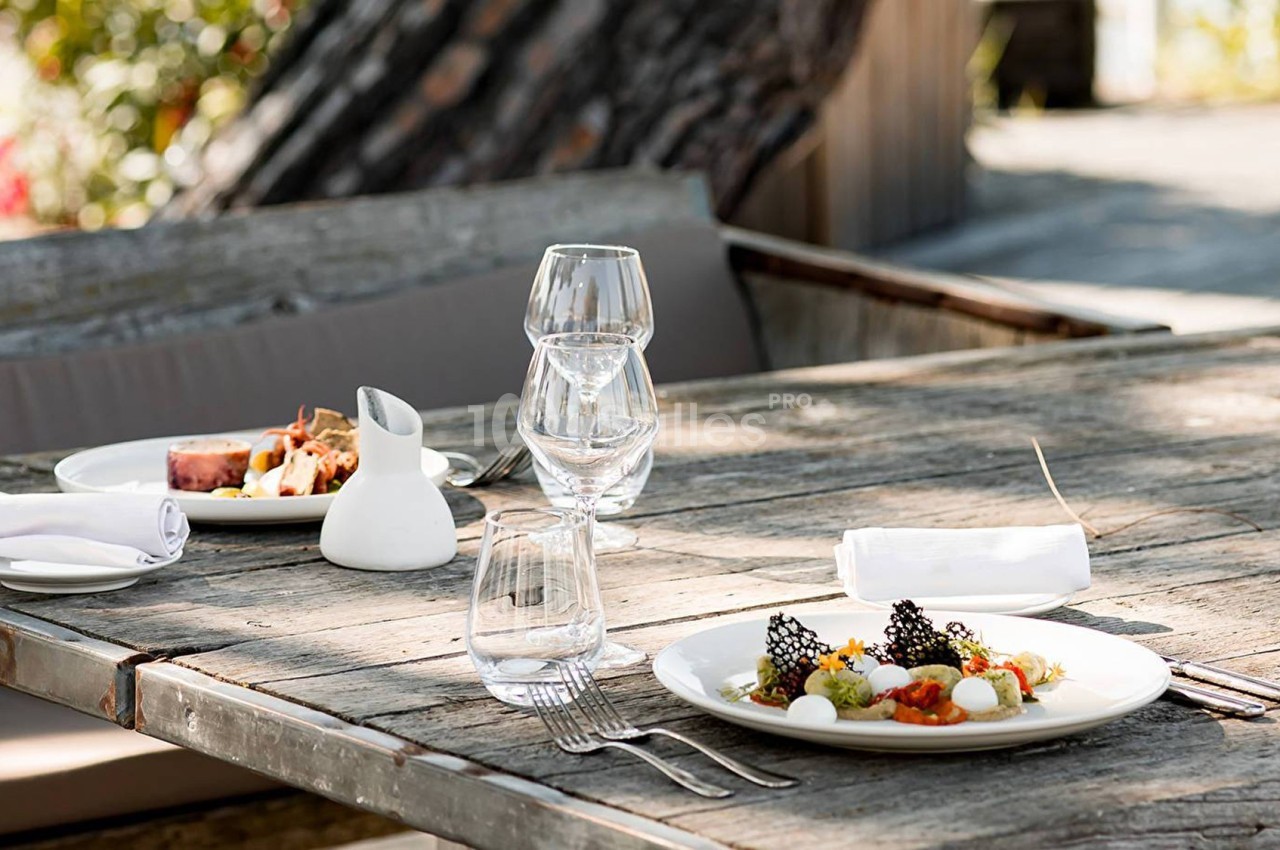 Table en bois avec deux assiettes de plats gastronomiques, verres et serviettes, en extérieur sous un arbre.