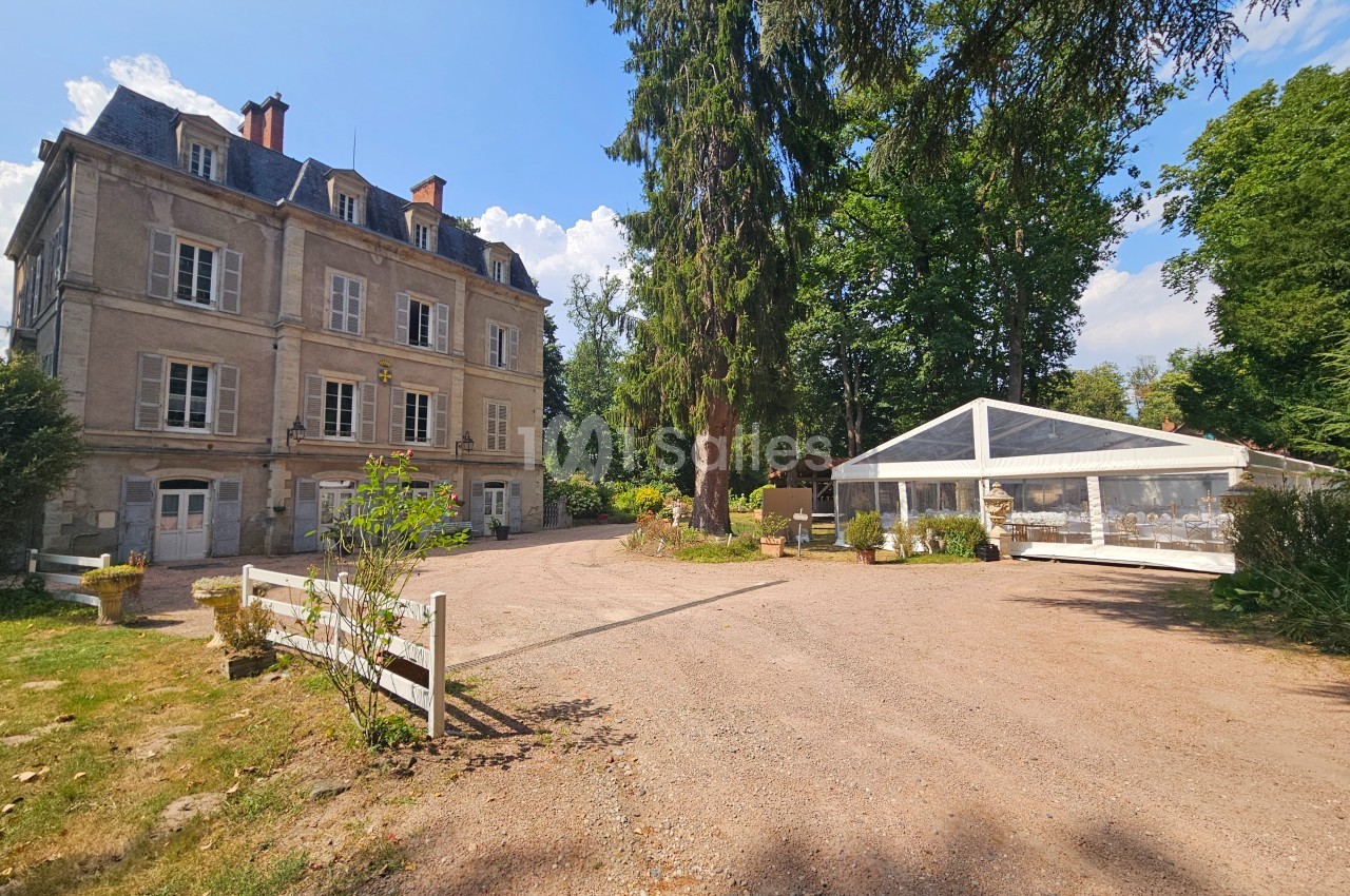 Façade d'un manoir ancien entouré d'arbres, avec une allée en gravier et une structure blanche attenante.