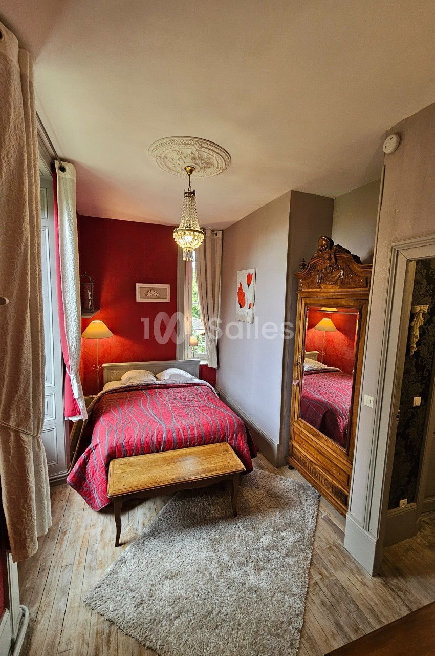 Chambre avec lit double, armoire en bois sculpté, lustre, murs rouges et gris, et tapis sur parquet clair.