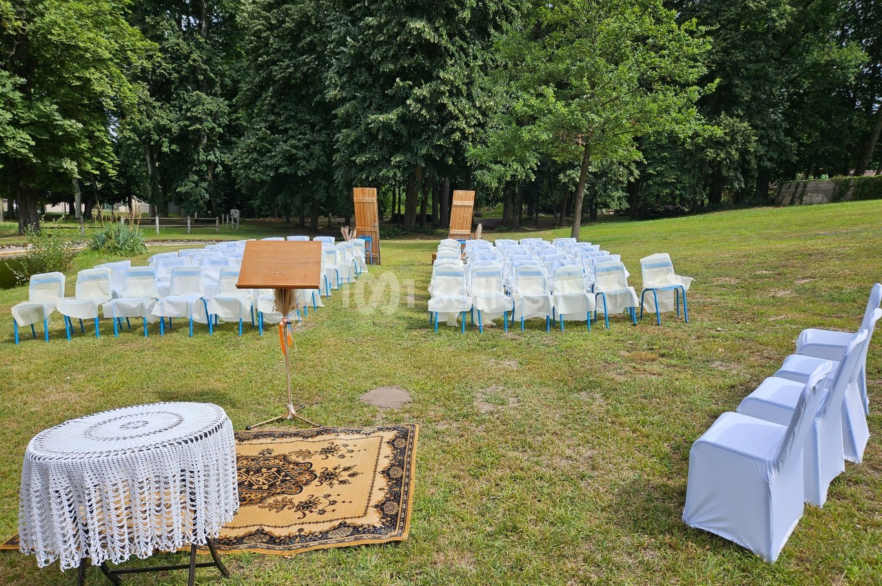 Chaises blanches disposées en extérieur sur une pelouse, face à un pupitre et deux portes décoratives en bois.
