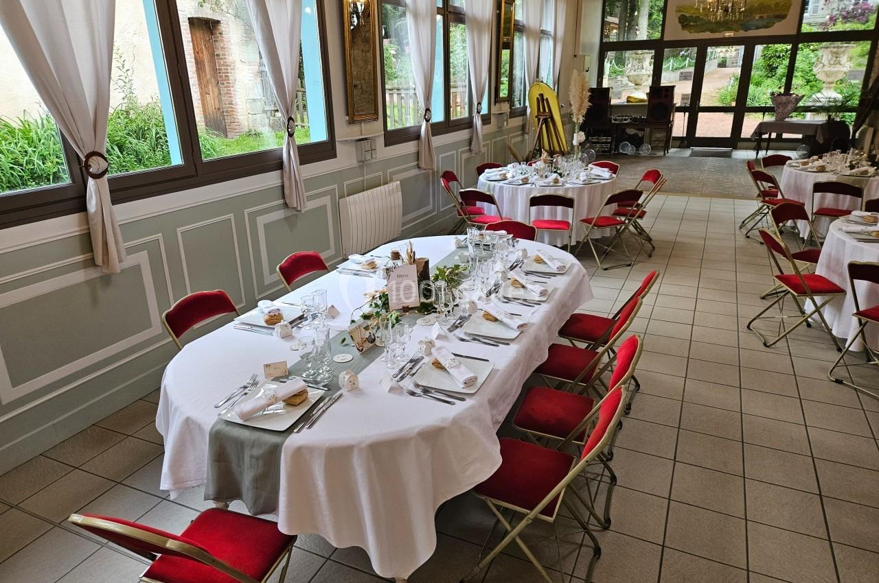 Salle de réception avec tables dressées, nappes blanches, chaises rouges et vue sur un jardin par des fenêtres.
