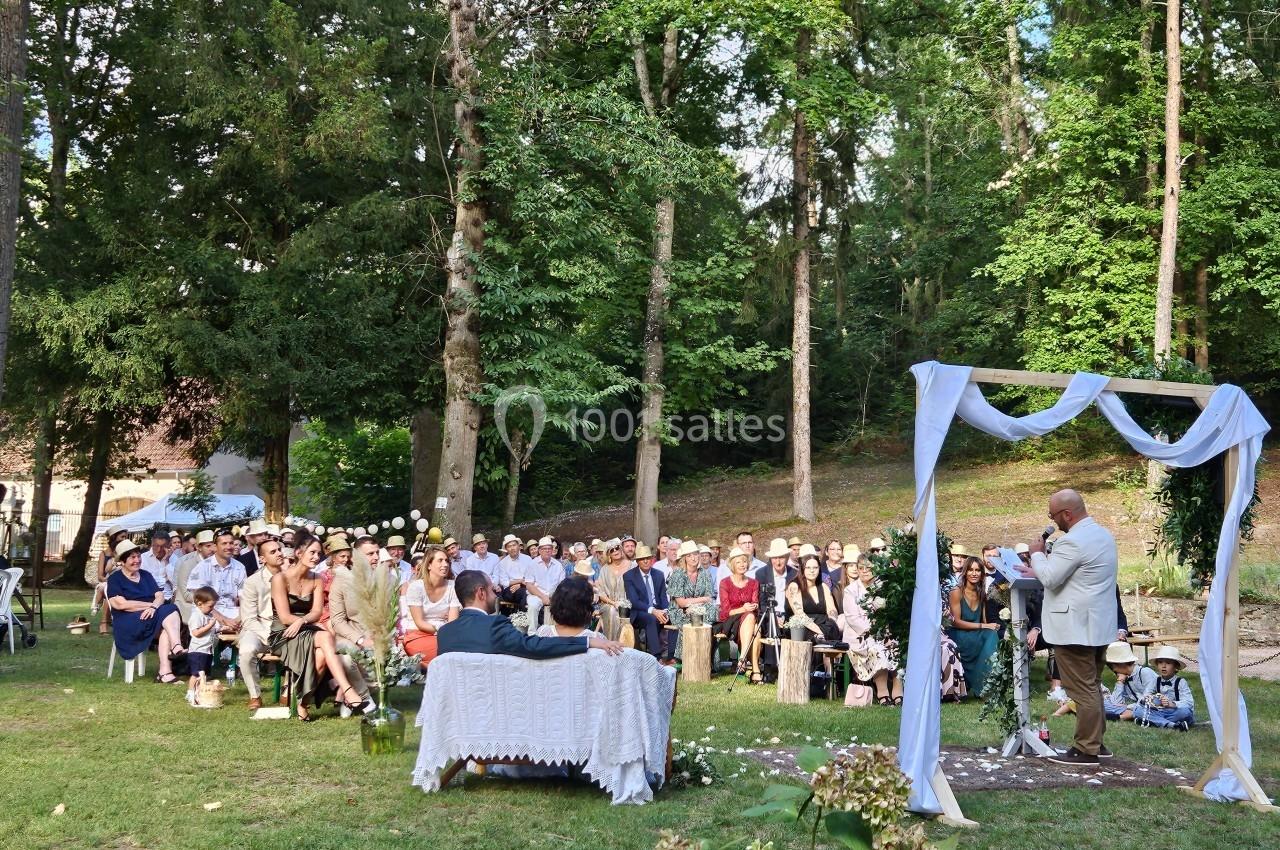 Cérémonie en plein air dans un jardin boisé, avec des invités assis et un officiant devant une arche décorée.