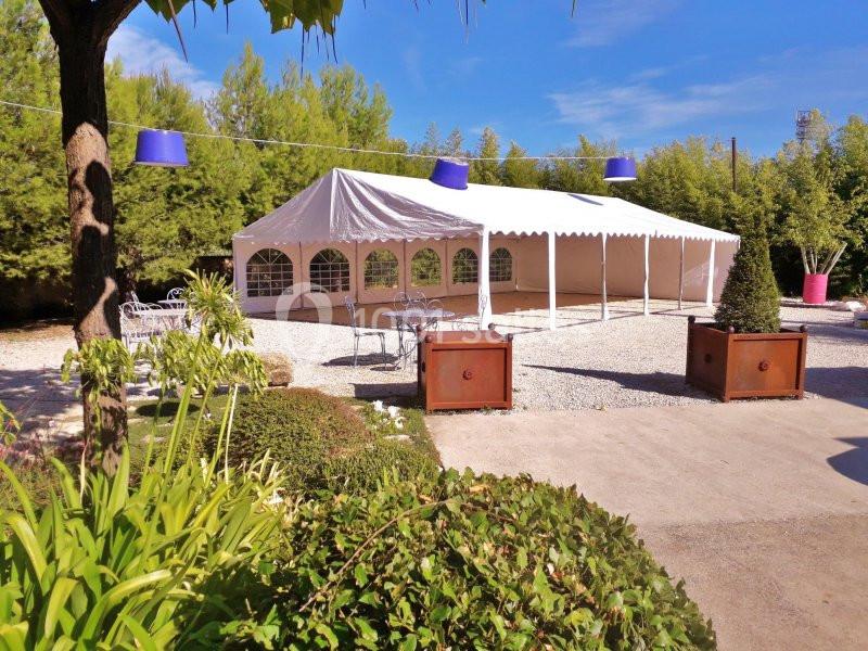 Tente blanche installée sur un sol gravillonné, entourée de végétation et de mobilier d'extérieur.
