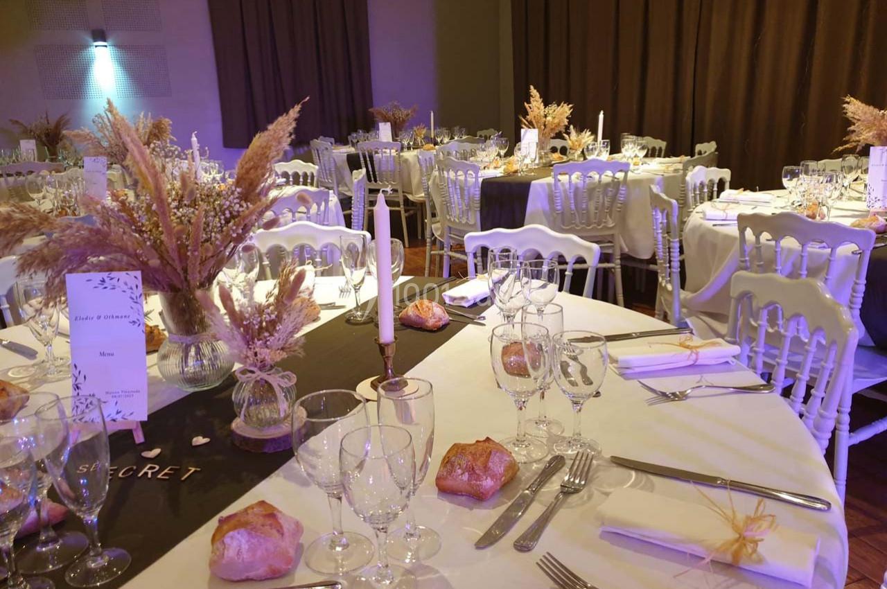 Salle décorée pour un événement avec tables rondes dressées, nappes blanches, bouquets de fleurs séchées et bougies.