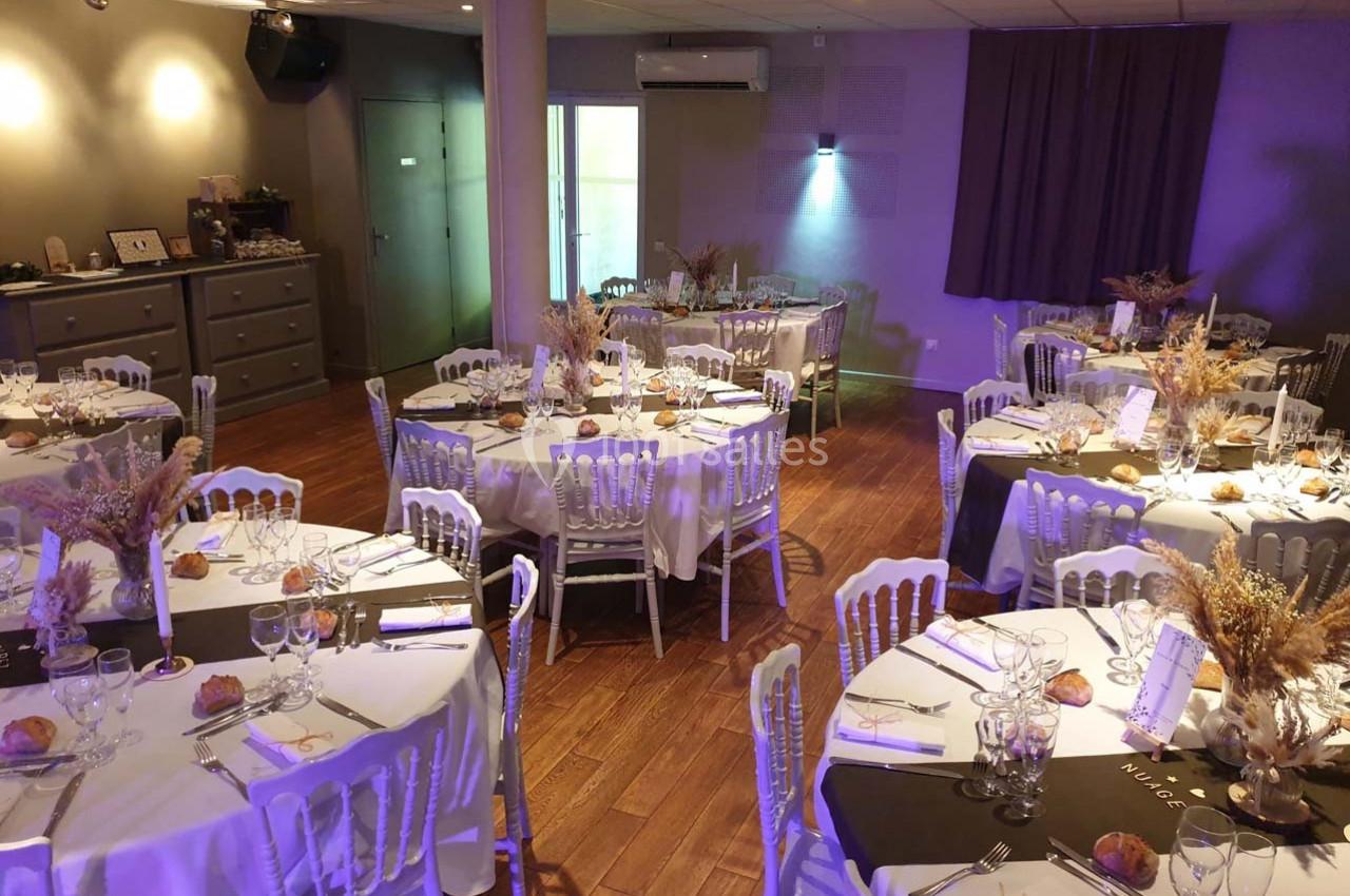 Salle de réception décorée avec des tables rondes dressées, nappes blanches, chaises blanches et éclairage tamisé.