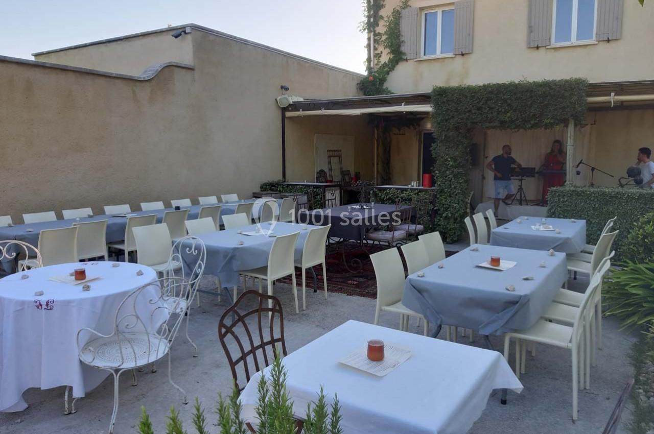 Terrasse extérieure avec tables dressées, chaises blanches, et musiciens jouant sous un auvent devant un bâtiment beige.