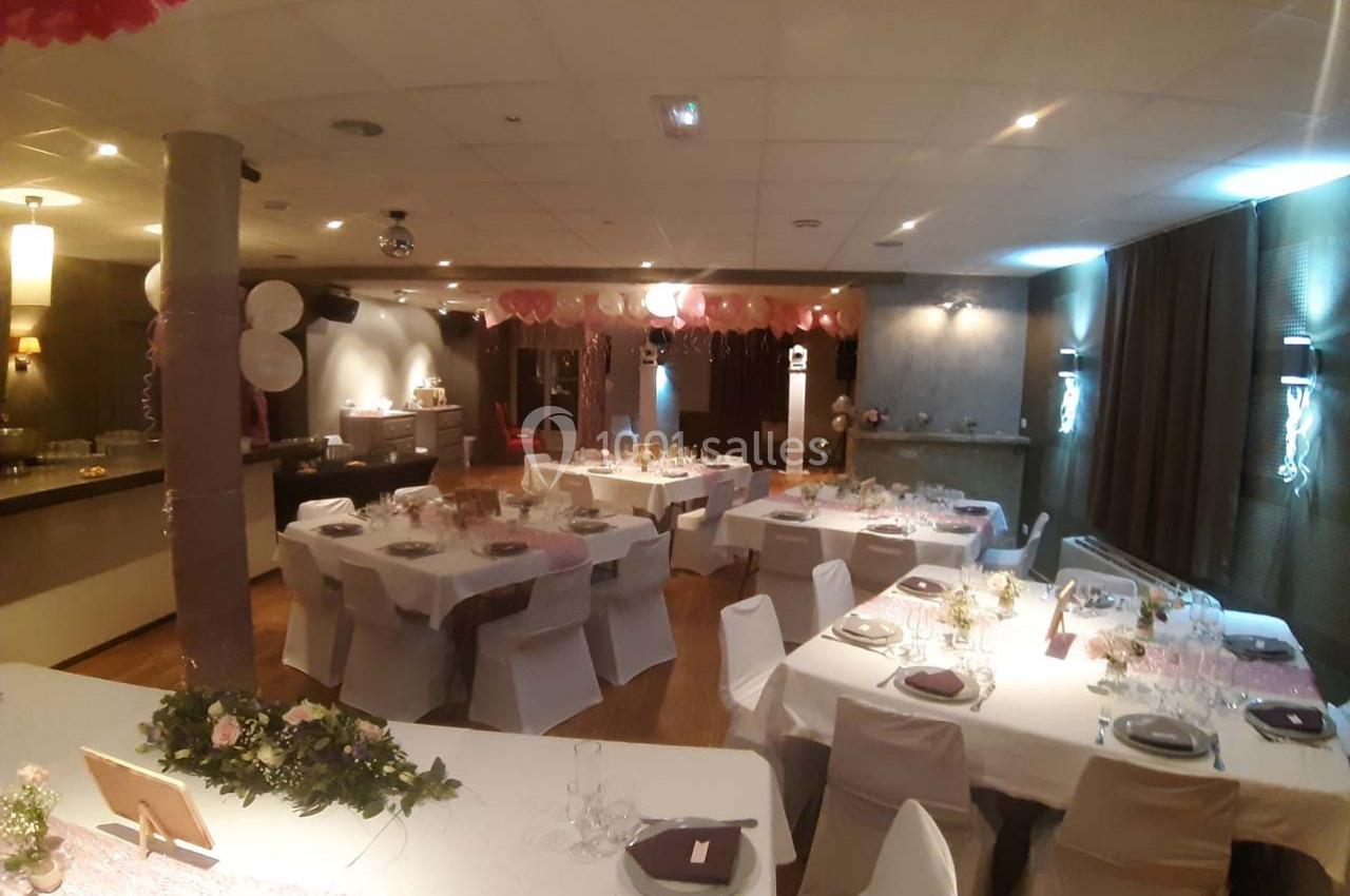 Salle de réception décorée avec des tables dressées, nappes blanches, décorations roses et ambiance lumineuse chaleureuse.