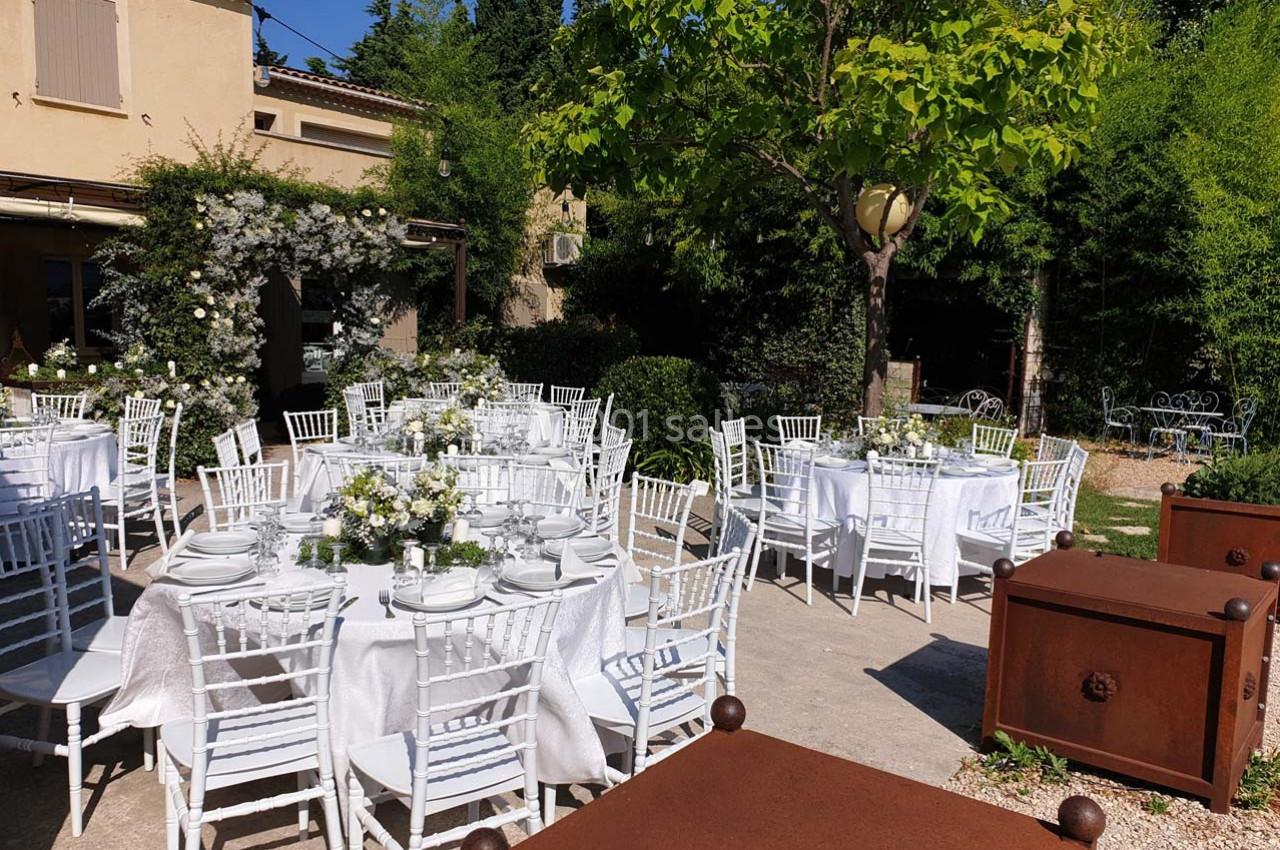 Tables rondes dressées avec nappes blanches et décorations florales dans un jardin ensoleillé près d'une maison.